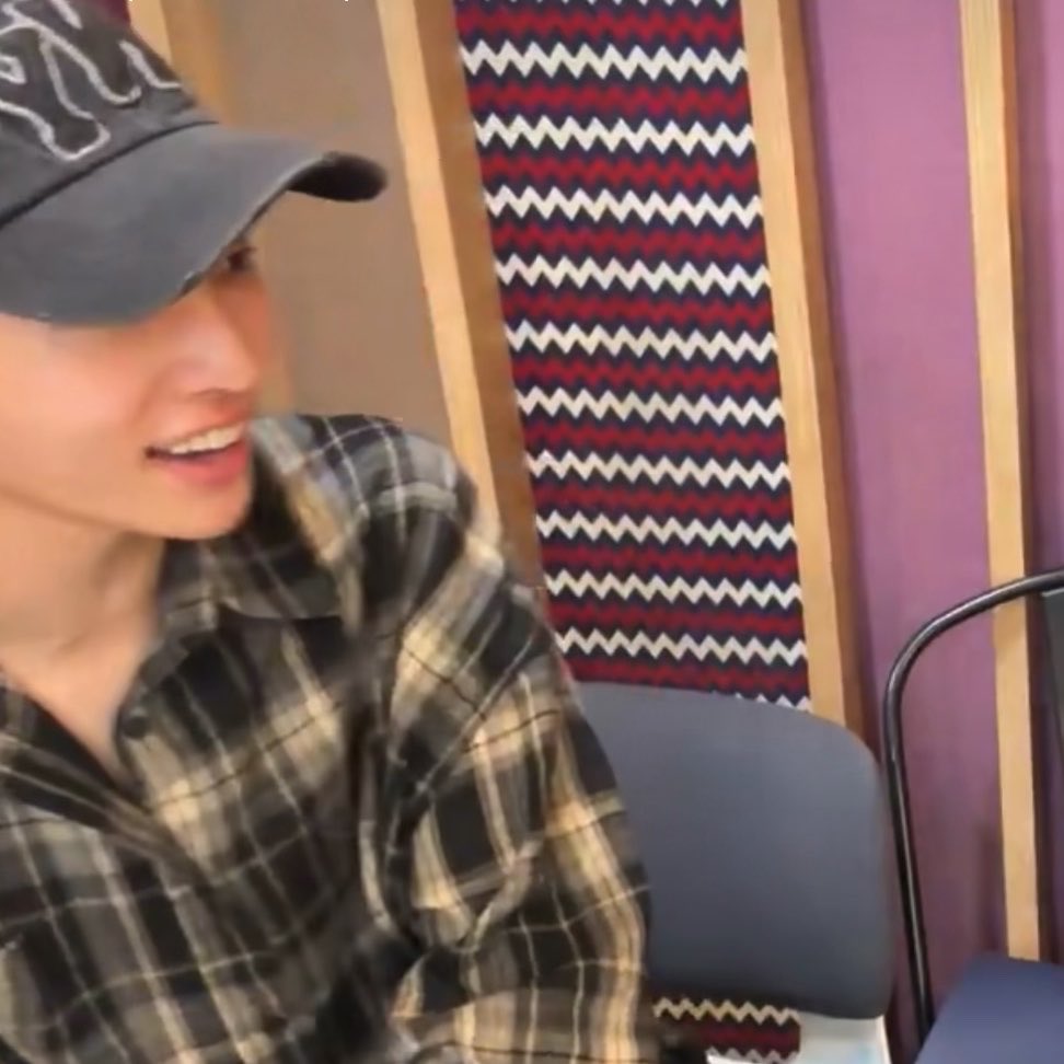 A side-profile shot of a smiling person wearing a dark baseball cap with white embroidery and a black and beige plaid flannel shirt, sitting indoors near a zigzag-patterned textile and a gray chair.