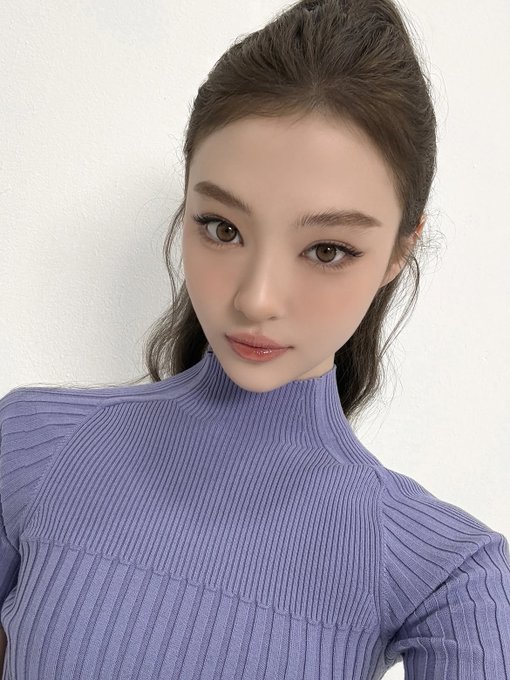 A young woman with dark hair styled in a high ponytail wears a ribbed lavender turtleneck sweater, looking softly at the camera against a plain white background.