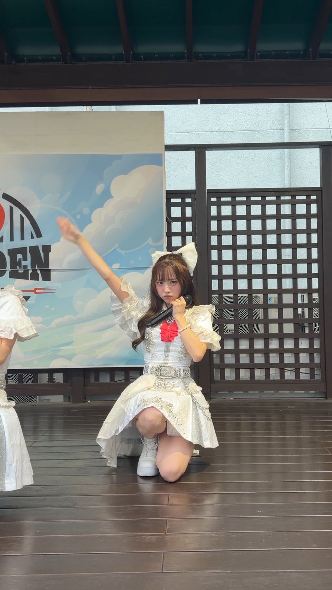 A young woman in a white dress and large bow performs on an outdoor stage with a wooden floor, holding a microphone while kneeling.