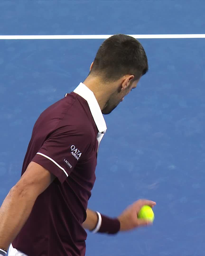 Tennis player Novak Djokovic, wearing a maroon "Qatar Airways" and "Lacoste" shirt, looks down while holding a yellow tennis ball on a blue court, likely preparing to serve during the China Open.