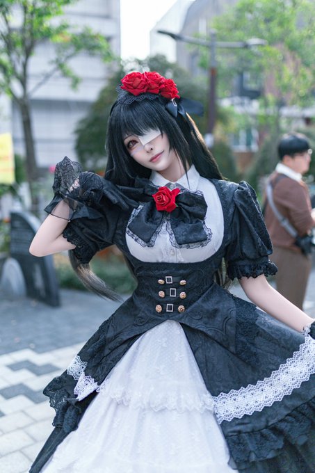 A cosplayer portrays Tokisaki Kurumi from "Date A Live," wearing a black gothic lolita gown adorned with red roses, a matching rose headpiece, and a distinctive eye patch over one eye, standing on an outdoor plaza.