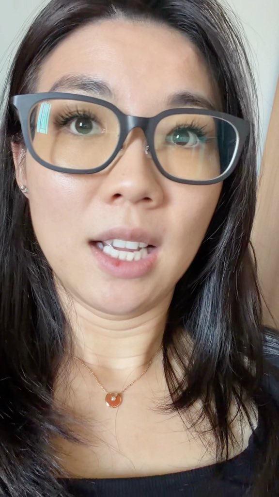 A close-up selfie-style photo of a woman with long dark hair wearing black-rimmed glasses, speaking with her mouth slightly open, and wearing an orange pendant necklace.
