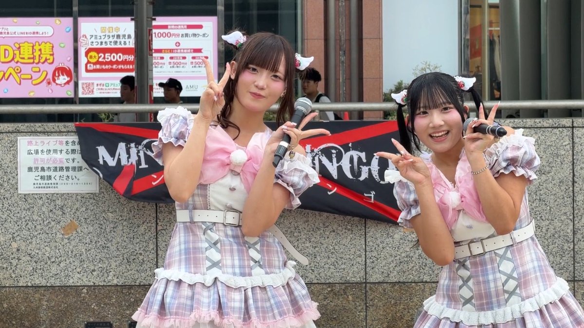 Two young women in matching pink and white plaid idol costumes with cat-ear headbands posing with peace signs and microphones outdoors, with Japanese promotional signage visible in the background.