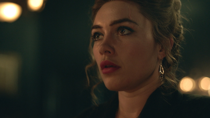 A woman with wavy hair and ornate earrings gazes off-screen with a somber expression, tear visible on her cheek, set against a dark background with warm bokeh lights. The image captures Florence Pugh in an emotionally charged moment as Cathy Ames in the upcoming Netflix adaptation "East of Eden."