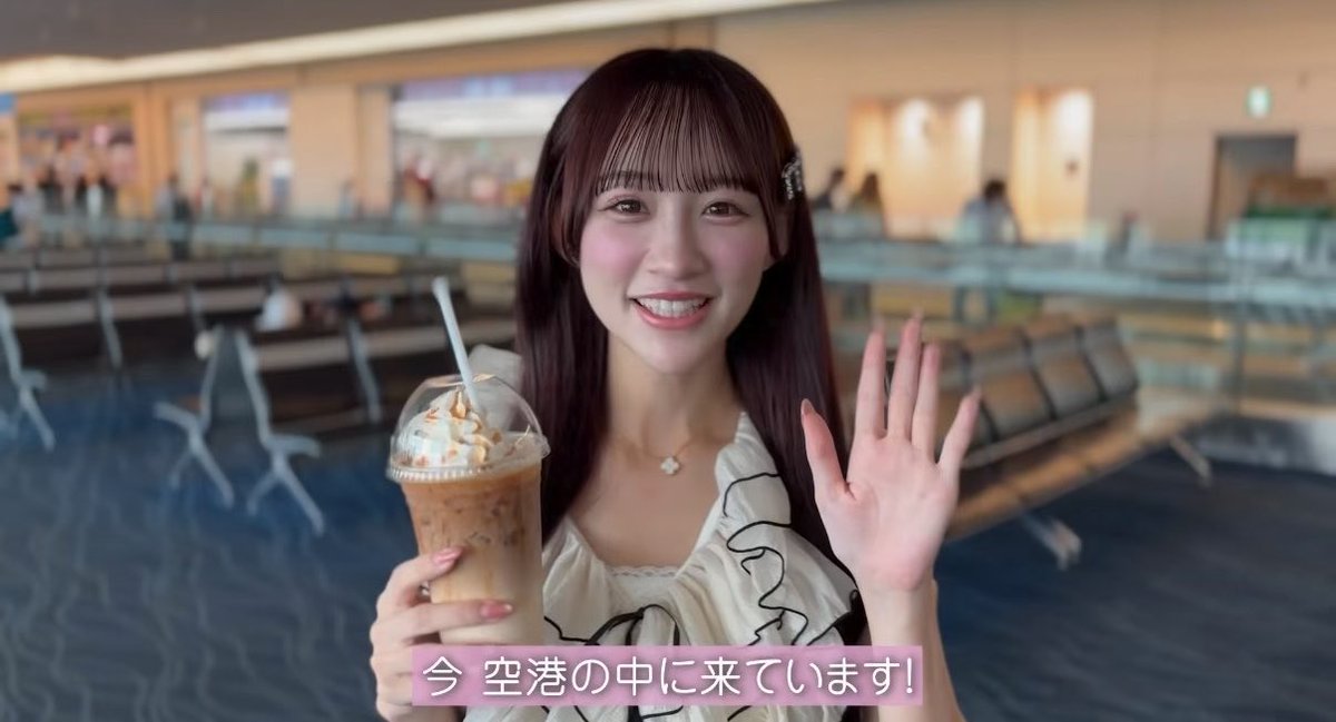 A young woman waves at the camera while holding an iced coffee topped with whipped cream in what appears to be an airport terminal, with Japanese subtitles reading "今 空港の中に来ています!" (Now I'm at the airport!). The cheerful, casual pose complements the lighthearted tone of the post.