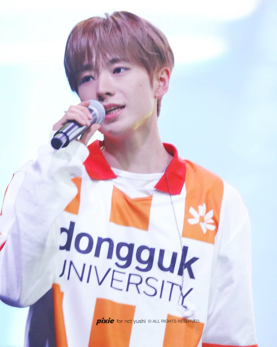 A performer with light brown hair sings into a microphone while wearing a white and orange jersey that reads "dongguk UNIVERSITY," with a watermark crediting the photo to "pixie for nct yushi," matching the post's reference to NCT WISH member YUSHI performing at Dongguk University.