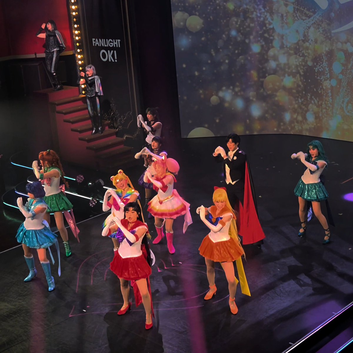 A live stage performance featuring performers dressed as Sailor Moon characters and Tuxedo Mask, with "FANLIGHT OK!" displayed on the screen behind them.
