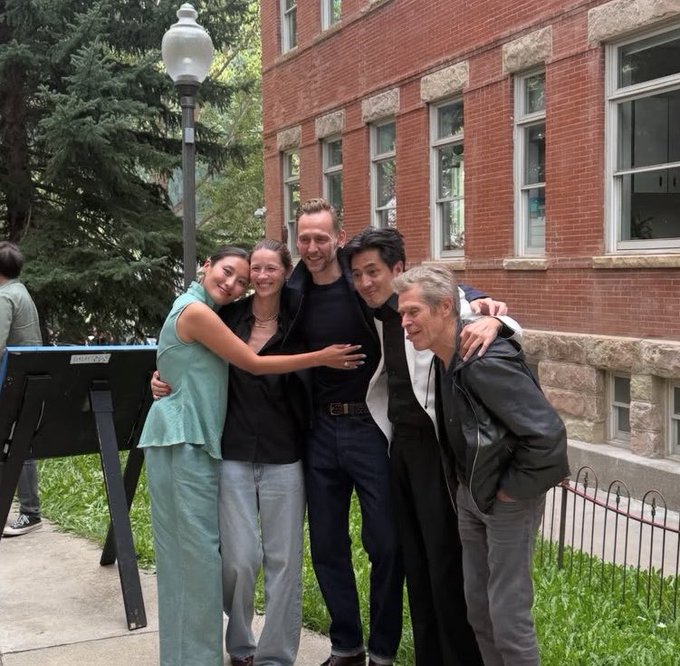 A group photo of five people smiling and posing with their arms around each other outdoors, standing next to a brick building and a street lamp.