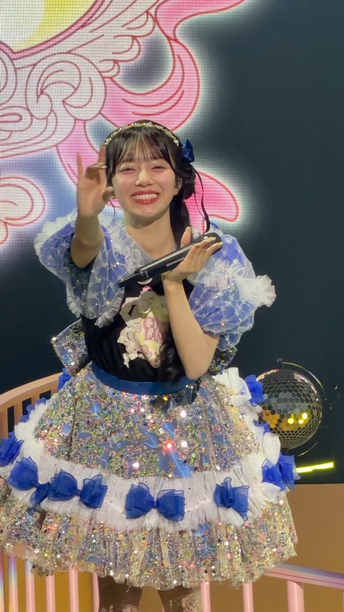 A young woman, likely a Japanese idol, wearing an elaborate blue and silver costume with ruffles and bows, smiling brightly and making a peace sign while holding a microphone on stage.