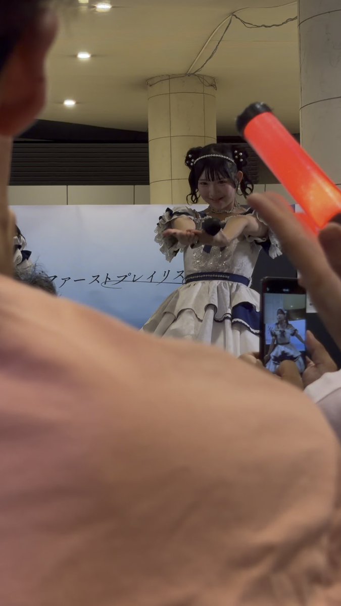 A performer in a blue and white frilly dress interacting with fans, while someone in the foreground holds up a red glow stick and a phone recording the event.