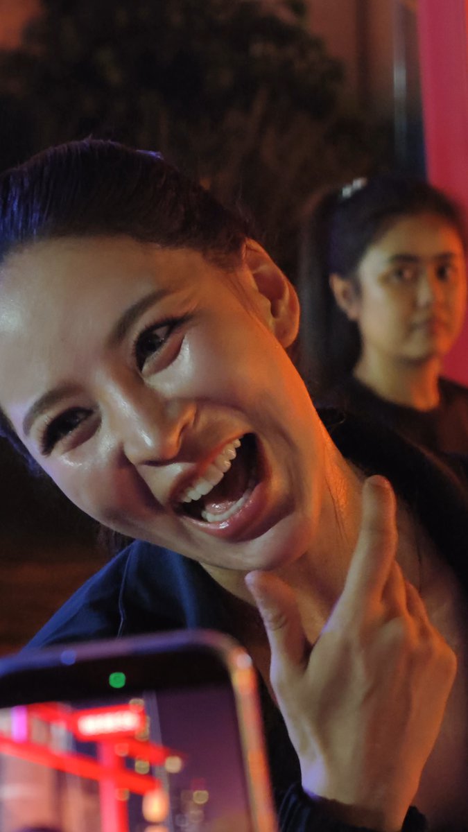 A woman laughs joyfully while making a peace sign, with a man visible behind her in a dimly lit, warm-toned setting.