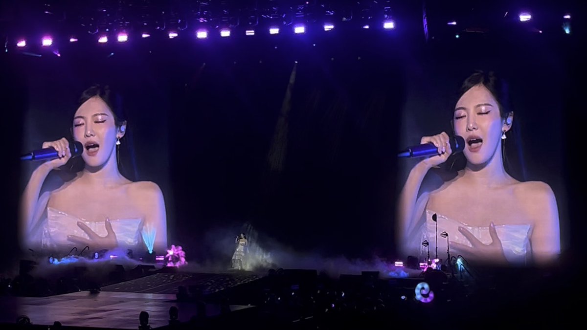 A concert scene showing a performer displayed on two large screens singing into a microphone, with stage lights and atmospheric effects visible.