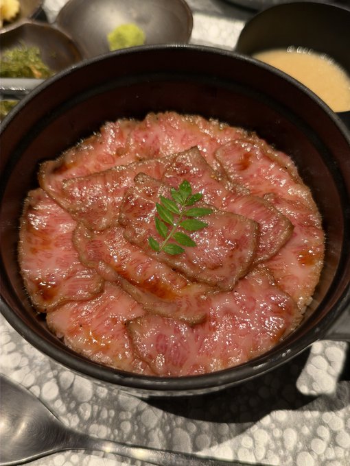 A black bowl filled with overlapping slices of pink, marbled Wagyu beef over rice, garnished with a sprig of green herbs, reflects the post's description of "Inoue" black Wagyu hitsumabushi, capturing the prized rare meat before it's mixed with dashi broth at the popular Ginza lunch spot.