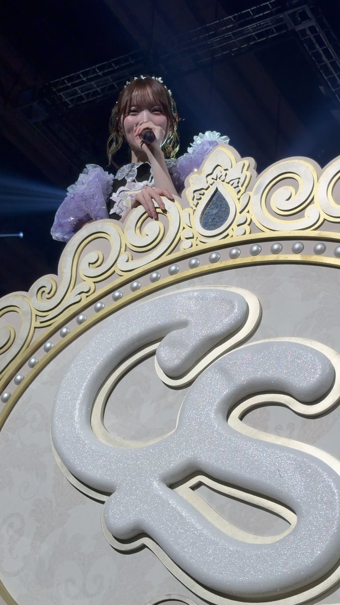 A female performer on stage singing into a microphone, wearing a purple and white frilly outfit with a tiara, positioned behind a large ornate gold and white decorative structure featuring a swirling symbol.