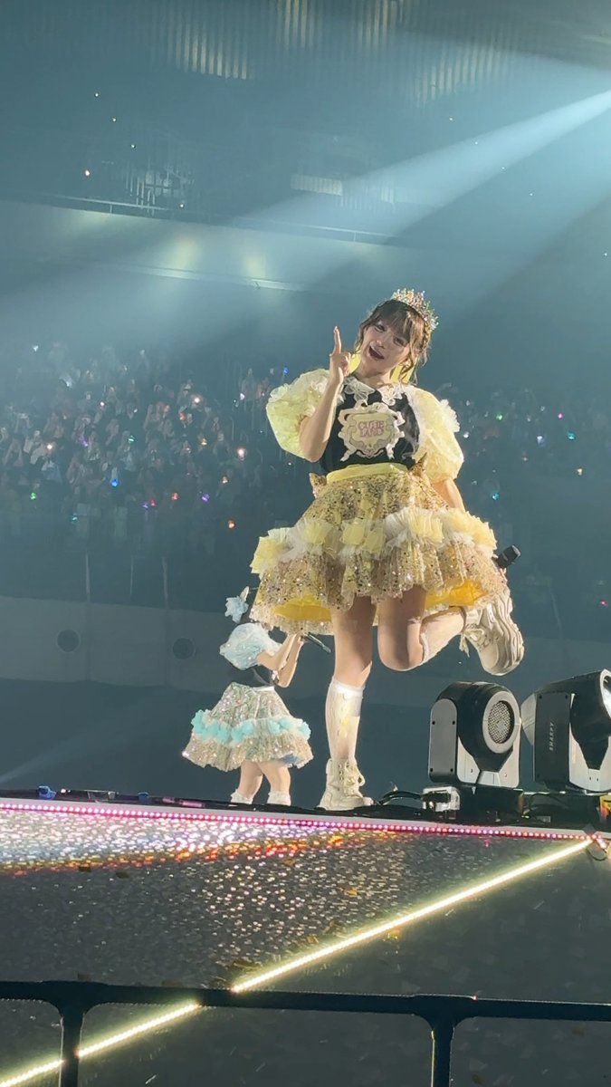 A female idol performer wearing a yellow and black frilly dress and a crown strikes a dynamic pose on a concert stage, with audience lights visible in the background.