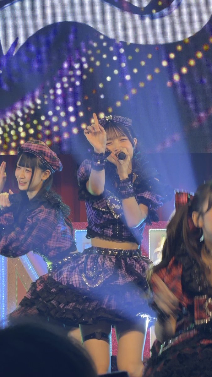 A performer in a purple plaid outfit sings into a microphone on stage with bright lights and a starry backdrop, flanked by other performers.