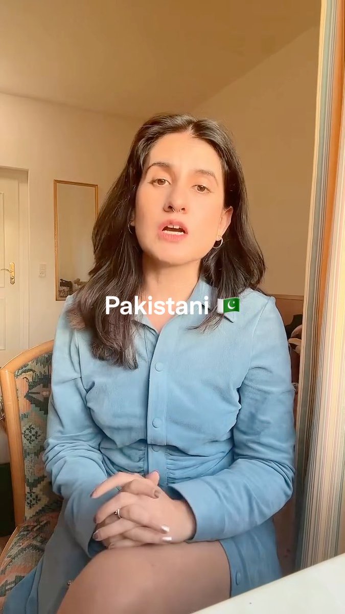 A woman with long dark hair and a nose ring, wearing a light blue button-up dress, sits indoors with a text overlay reading "Pakistani 🇵🇰".