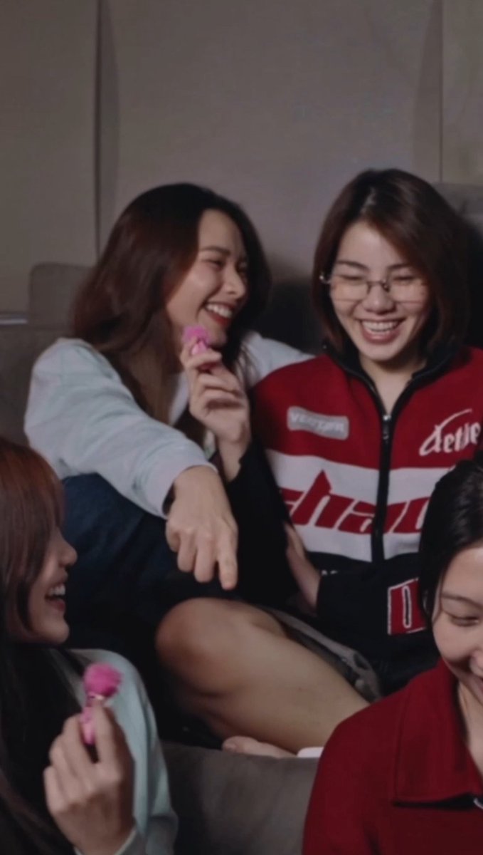Three young women laugh together on a gray couch; one wears a red and white jacket with 'dev' visible on it, while another holds up a small pink object.