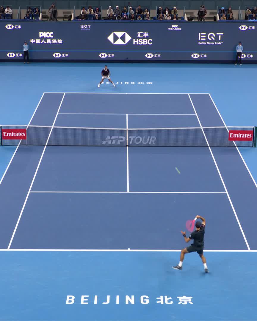 A tennis match at the China Open in Beijing features Djokovic (#DjokerNole) mid-serve at the baseline while his opponent prepares to return from across the net, with HSBC and ATP Tour signage visible around the blue court.