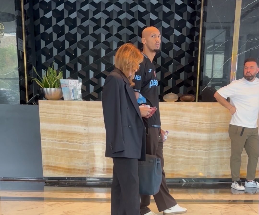A scene in what appears to be a hotel lobby where a woman in a dark blazer walks past a man in a black jersey, with another man in a white shirt standing by the reception desk.