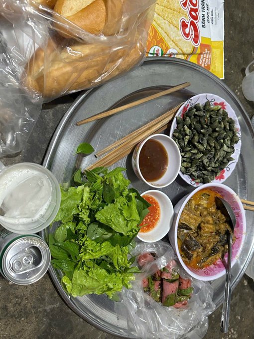 A metal tray holding Vietnamese-style dishes, including fresh herbs, dipping sauces, what appears to be stir-fried insects, and bread, alongside a can of beverage.