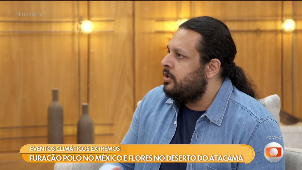 Screenshot from Brazilian TV show 'Encontro' showing a bearded man in a denim shirt, with text overlay about extreme weather events including 'FURACÃO POLO NO MÉXICO E FLORES NO DESERTO DO ATACAMA' and 'AO VIVO' indicator.
