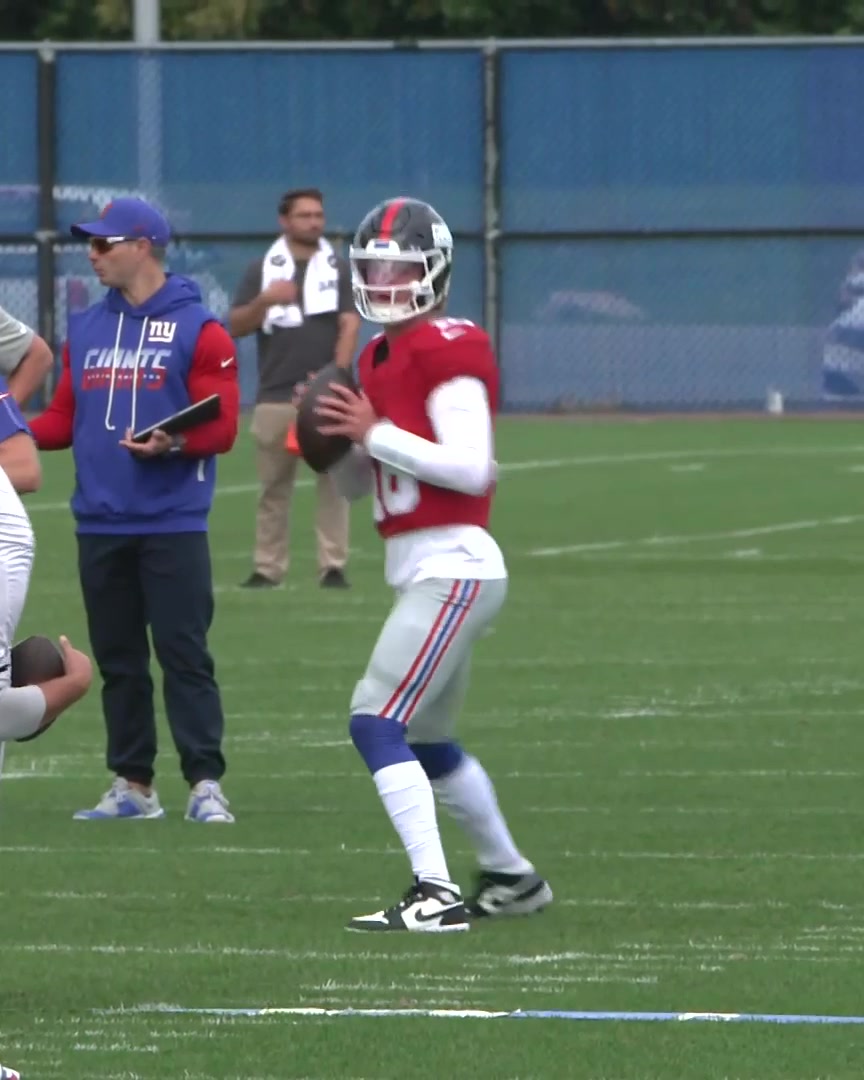 A quarterback wearing a red No. 18 jersey and helmet prepares to throw a pass at practice, while a coach in a blue hoodie with "NY GIANTS" branding observes on the sideline. The scene aligns with the post showing J.J. McCarthy in his Giants uniform during a training session.