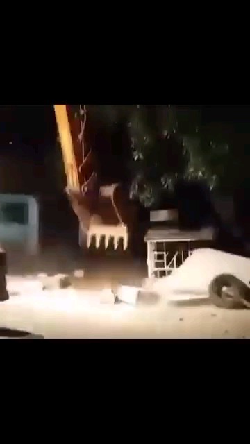A dark, blurry nighttime scene showing an excavator with its bucket lowered near scattered concrete blocks and debris on a dirt surface.