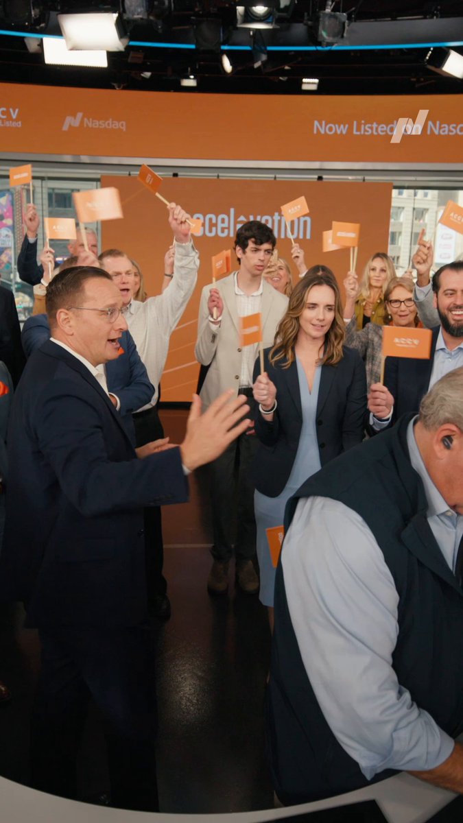 A jubilant crowd celebrates Accelevation's Nasdaq listing at the exchange, with attendees waving orange "$ACCV" flags beneath "Now Listed on Nasdaq" signage, capturing the company's milestone market debut.