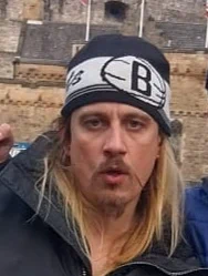 A close-up portrait of a man with long blonde hair and a moustache, wearing a black and white beanie.