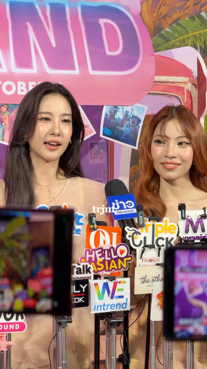 Two young women stand behind a wall of media microphones at a press event, with visible outlet signs including 'trending', 'Triple Click', 'Hello Asian', 'WE intrend', 'the dsa', and a backdrop with 'AND' and 'GLOBAL'.