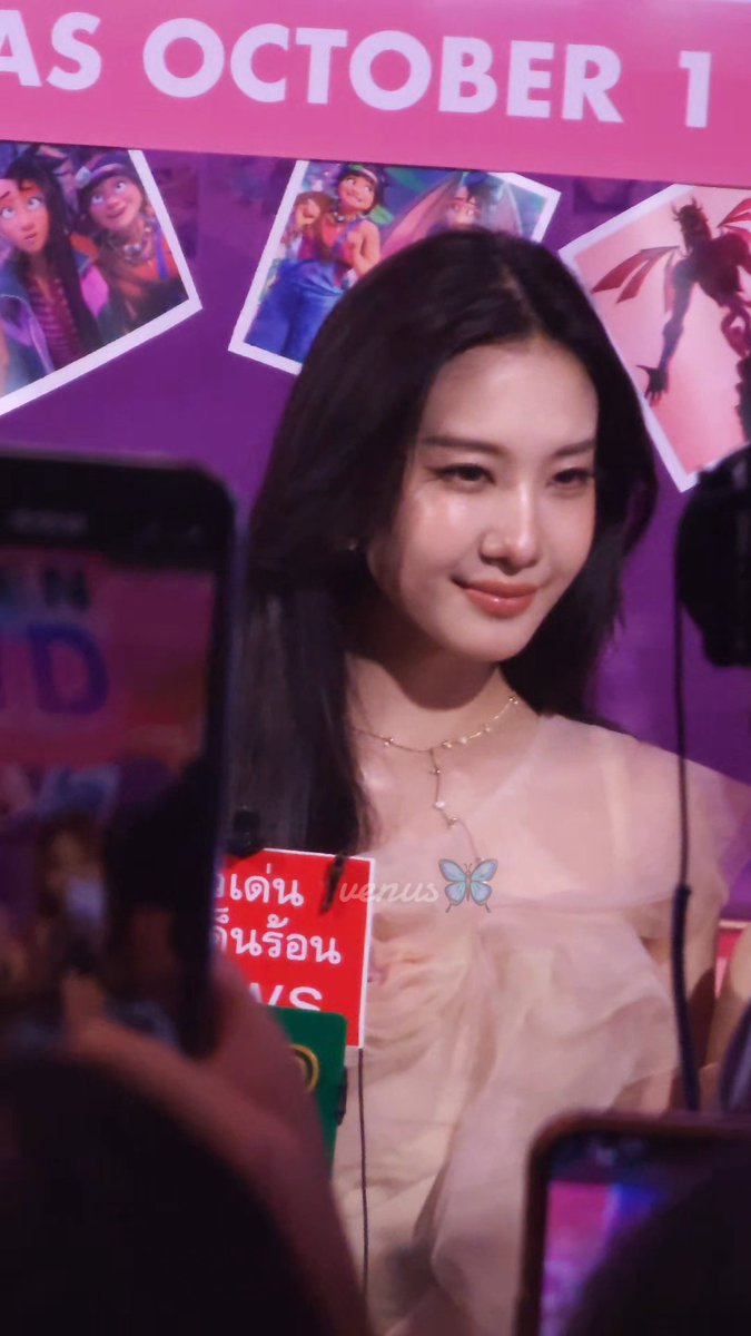 A woman at a press event smiles in front of a pink banner reading 'AS OCTOBER 1' with 'venus' and a butterfly logo visible.
