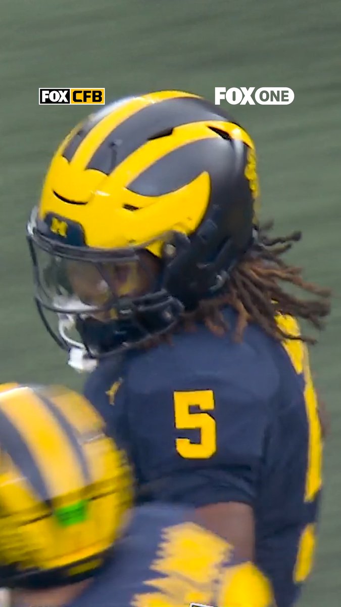 A Michigan Wolverines football player wearing a yellow and blue helmet with dreadlocks, with FOX CFB and FOX ONE logos displayed.
