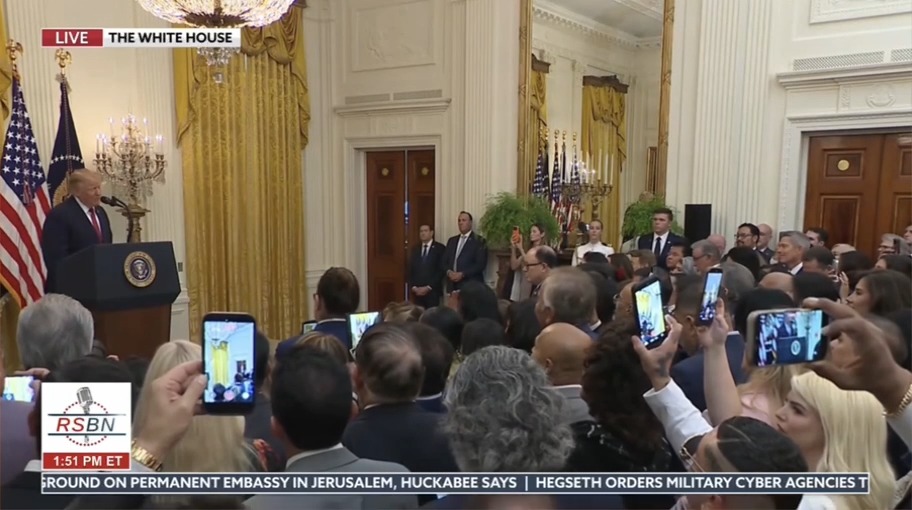 A screenshot of an RSBN live broadcast from the White House shows President Donald Trump speaking at a podium to a packed East Room audience, many holding up phones to record, with a news ticker mentioning Huckabee's comments on a permanent embassy in Jerusalem and Hegseth's military cyber agencies order, while a separate caption notes "Diesel is $6.39 today"—the unrelated text appearing to comment on fuel prices despite the political backdrop.