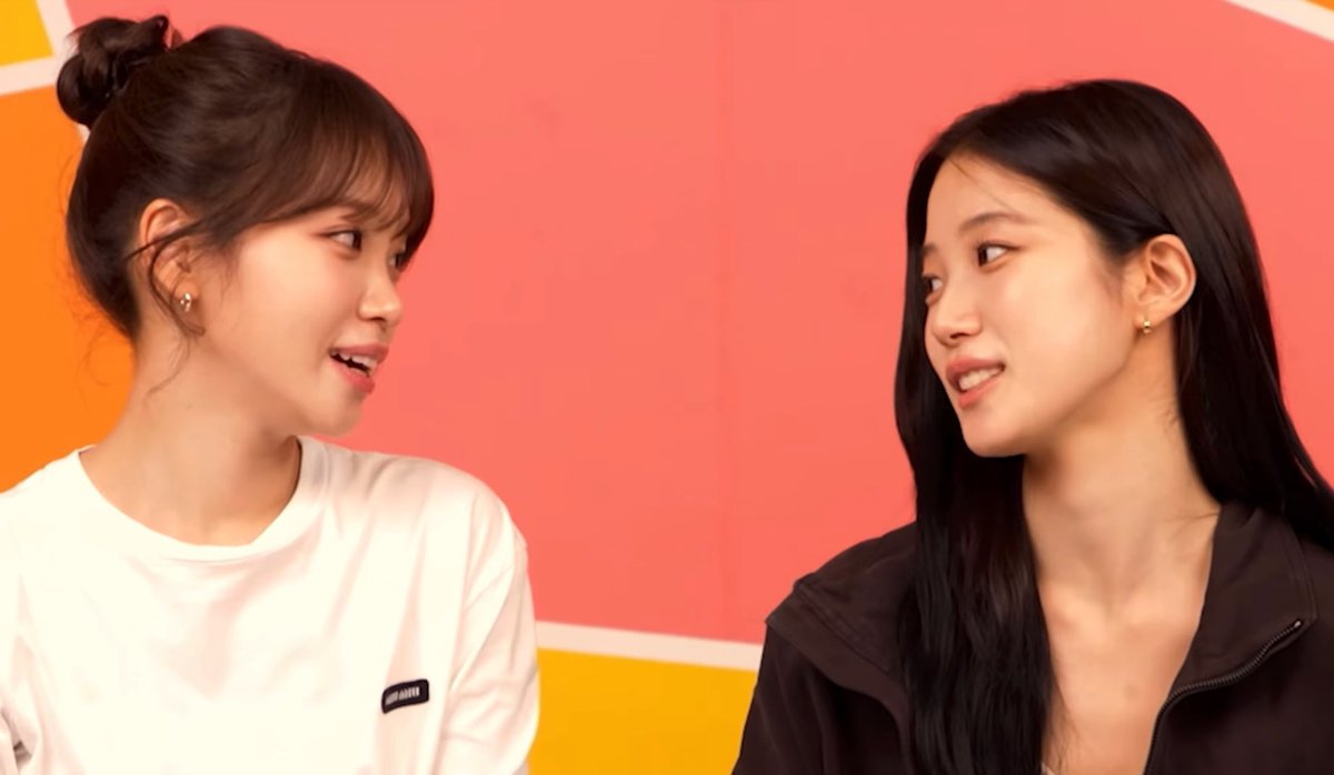 A photograph of two young women facing each other in conversation against a bright orange and yellow background.