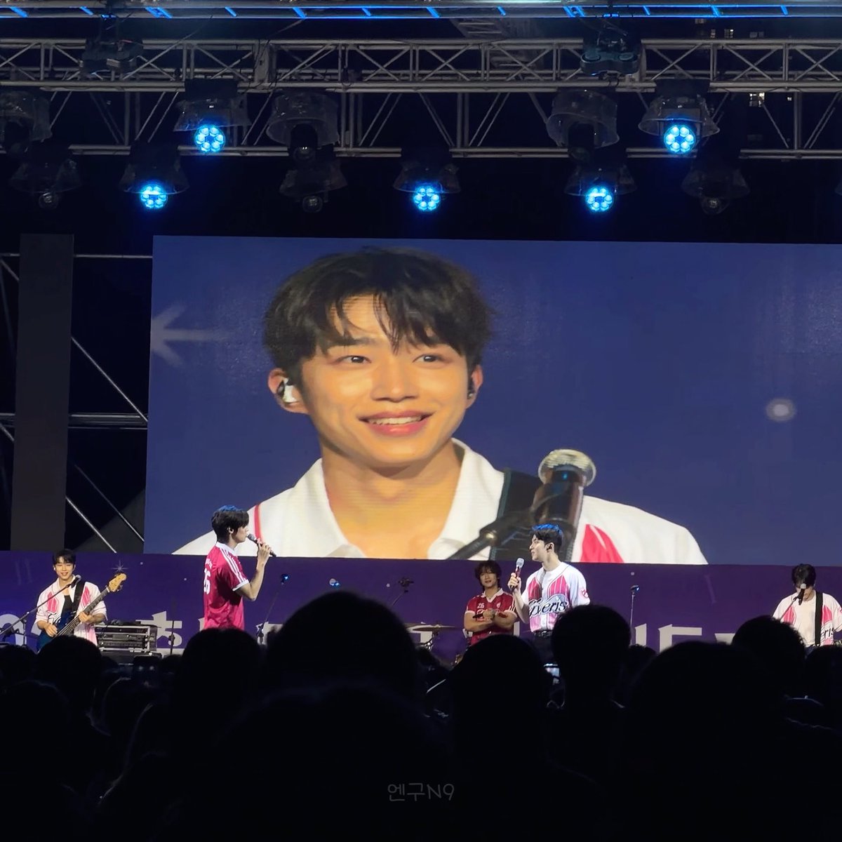 A concert scene featuring a large screen displaying a smiling young man's face, with band members visible below on stage and an audience silhouette in the foreground.