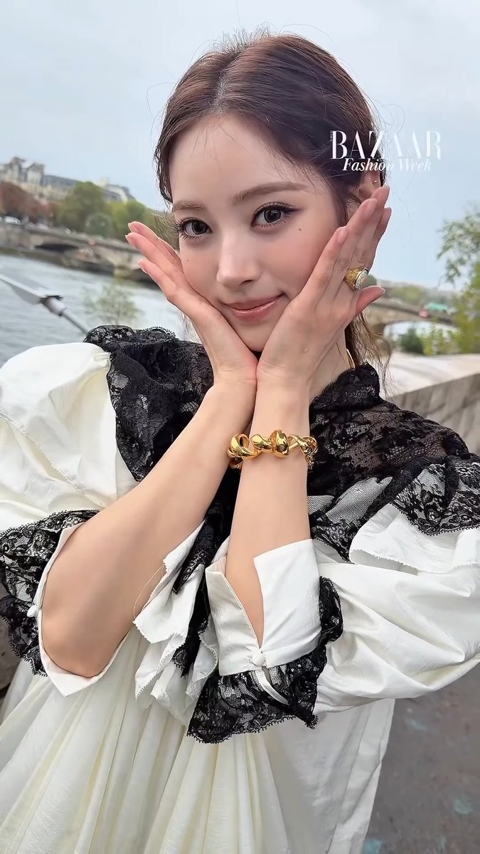 Selfie of a woman posing with her hands framing her face, wearing a white blouse with black lace details and a gold bracelet, with "BAZAAR Fashion Week" text overlaid.