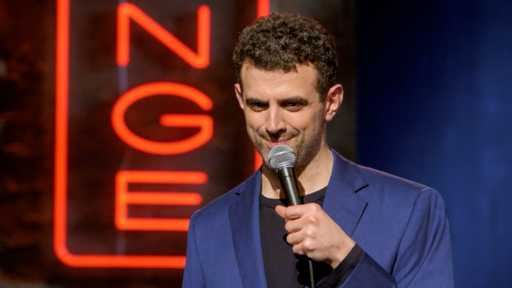 A man in a blue suit holds a microphone while performing on stage in front of a red neon sign.