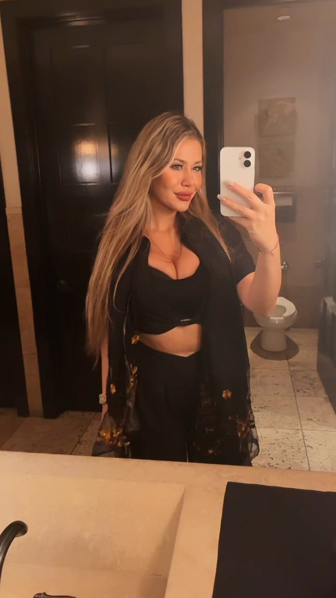 A blonde woman takes a mirror selfie in a bathroom wearing a black crop top and a sheer black cover-up with floral patterns.
