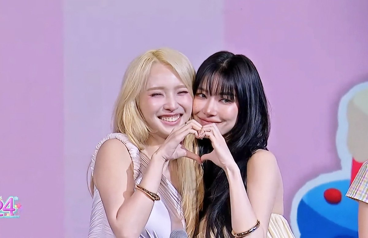 Two young women, one with blonde hair and one with dark hair, smiling warmly and forming a heart shape with their hands together at a birthday event.