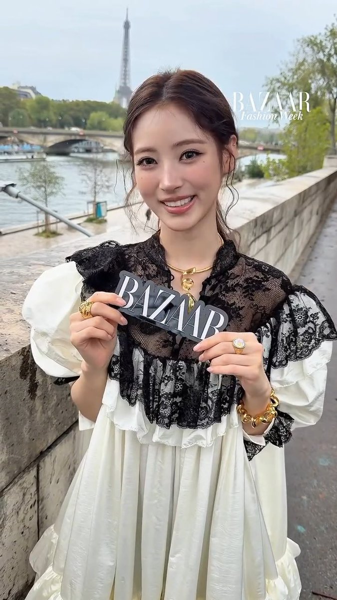A woman stands on a Parisian bridge with the Eiffel Tower in the background, holding a 'BAZAAR' sign; a 'BAZAAR Fashion Week' watermark appears in the upper right.