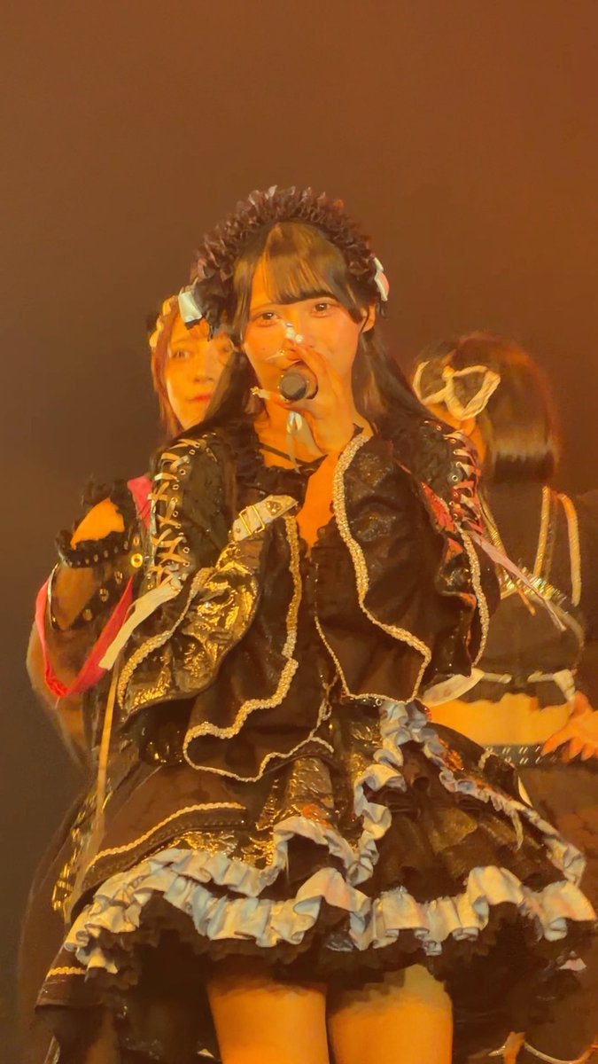 A young woman in an elaborate black gothic-style costume with lace, ruffles, and gold trim sings into a microphone on a dimly lit stage, with another performer visible behind her.