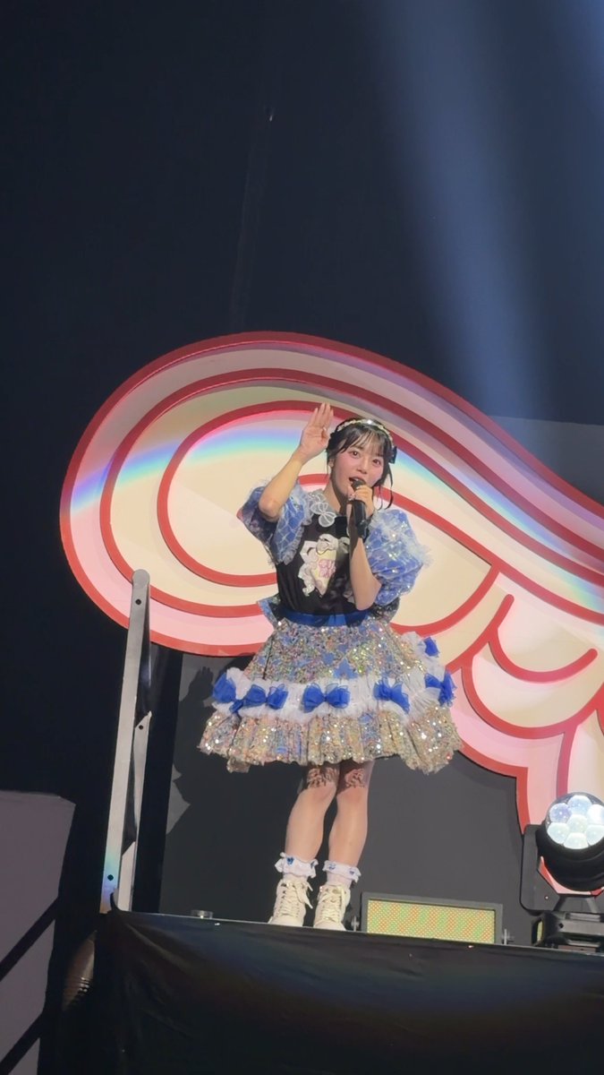 A young woman wearing a blue and white frilly idol-style dress performs on stage, holding a microphone with a colorful swirled backdrop behind her.