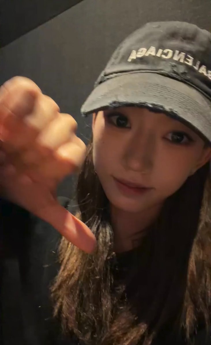 A selfie of a young woman wearing a distressed gray 'BALENCIAGA' cap, giving a thumbs down to the camera.