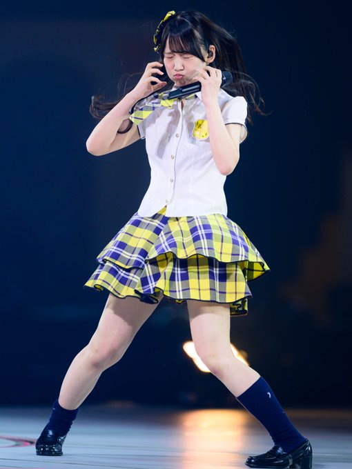 A performer on stage is captured in a dynamic pose, wearing a white short-sleeved blouse, a yellow and purple plaid skirt, and dark knee-high socks, holding a microphone.