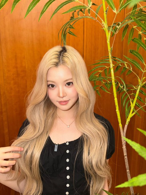 A young woman with long, wavy blonde hair and a black button-up top poses in front of a wooden wall next to a green plant.