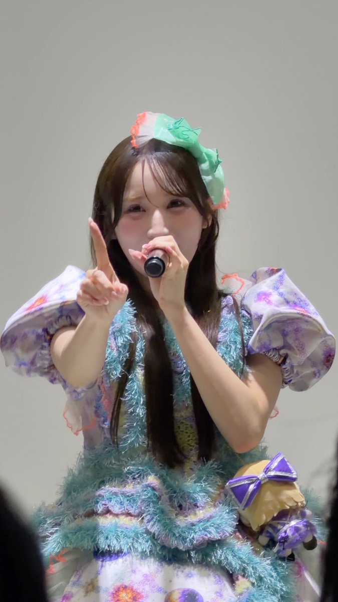 A young performer wearing a colorful pastel outfit with puffy sleeves and a green headband holds a microphone and points upward while on stage.