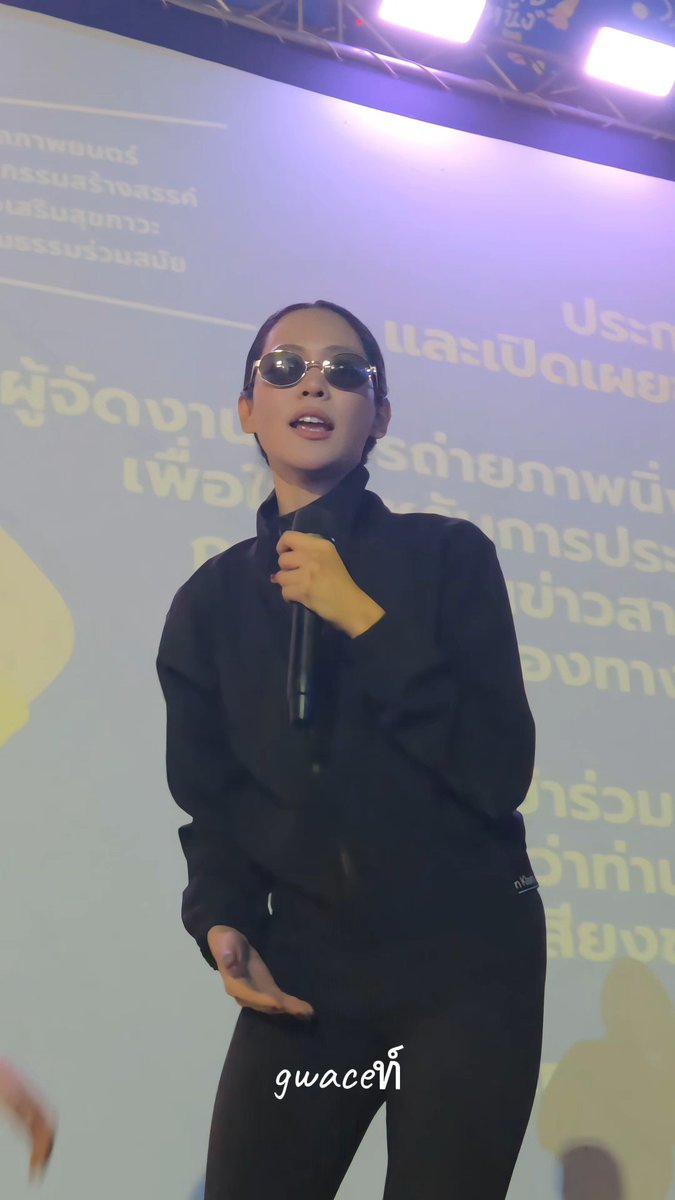 A woman with short hair wearing sunglasses and a black zip-up jacket holds a microphone while standing in front of a screen with Thai text and the watermark 'gwaceที'.