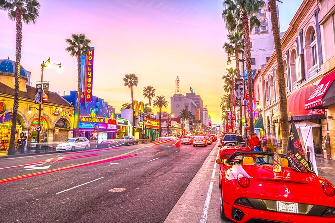 A vibrant sunset street scene in Hollywood featuring the iconic 'HOLLYWOOD' sign on the El Capitan Theatre, a Dolby Theatre marquee, palm trees lining the street, light trails from traffic, and a red convertible Ferrari parked in the foreground.