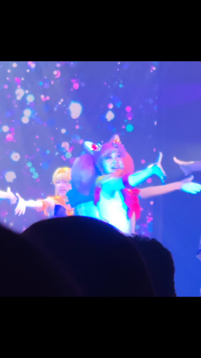 A performer on stage with orange twin-tail hair and a red bow extends their arms outward in a theatrical pose, illuminated by colorful bubble-like stage lights.